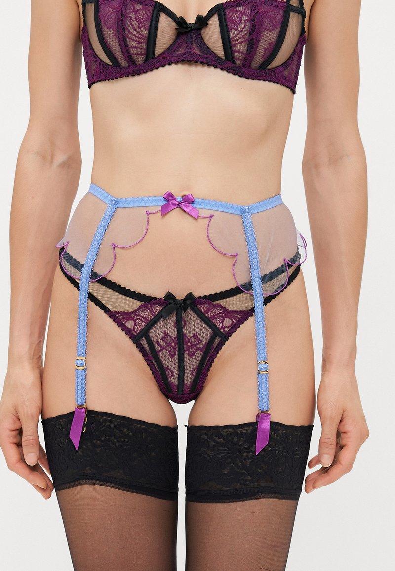 Donna che indossa un reggiseno di pizzo viola e uno slip coordinato, con una cintura per calze trasparente blu e calze autoreggenti in pizzo nero.