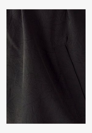 Tessuto nero con una superficie testurizzata, dotato di una tasca laterale e lievi pieghe. Il materiale appare leggero e morbido.