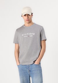 Siva pamučna majica s okruglim izrezom i kratkim rukavima, s bijelim natpisom "HILFIGER USA 1985". Uparena s svijetloplavim trapericama i bež kapom.