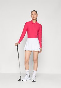 Ružová dlhá rukávová polo košeľa, biela rozšírená golfová sukňa, biele tenisky a golfová hůl. Oblečenie má na sukni logo Nike.