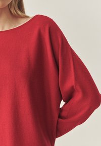 Pull en tricot rouge avec un col bateau et des manches amples. La texture semble douce et lisse, avec une couleur unie sans motifs.