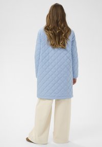 Svetlo modra prešita dolga jakna z vzorcem diamantov, kombinirana s kremnimi hlačami širokih hlačnic in rjavimi čevlji. Gladek tekstura, praktična zasnova.