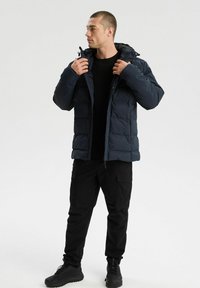 Mężczyzna dostosowujący kaptur w granatowej kurtce puffer, noszący czarne spodnie cargo, czarną koszulę i czarne sneakersy, stojący na białym tle.