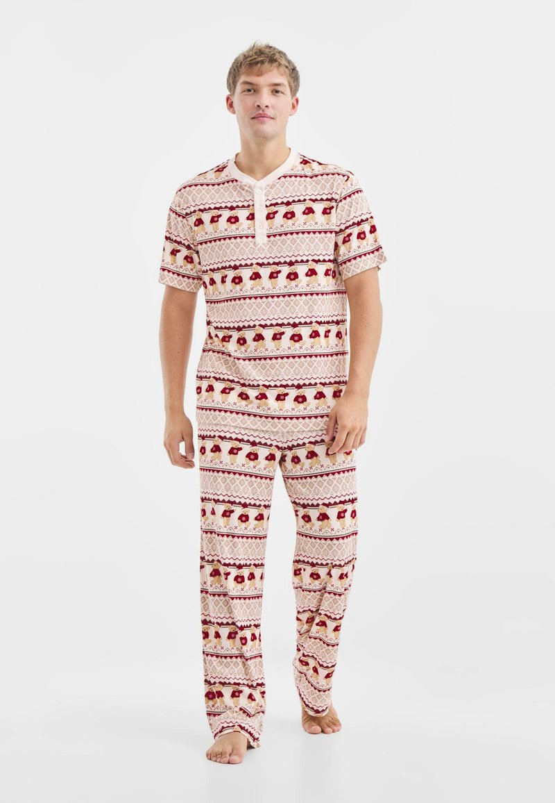 Lange pyjamaset in beige met een rood en groen patroon. Heeft korte mouwen, een gebuttoneerd kraagje en loszittende broekspijpen.
