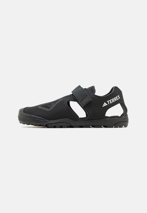 Zapatilla deportiva negra con parte superior de malla, diseño abierto y correa ajustable. Suela texturizada para tracción. Logo de la marca visible.