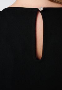 Zwarte jurk met een keyhole-ontwerp op de rug, afgesloten met een decoratieve ronde knoop. Gemaakt van een soepele stof met een subtiele textuur.