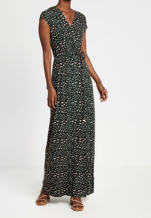 Vrouw draagt een zwarte korte mouwen overslag maxi-jurk met een klein bloemenprintje en beige sandalen met bandjes.