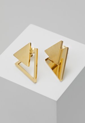 Paire de boucles d’oreilles géométriques en or avec des formes de triangles pleins et ouverts exposées sur une surface blanche.