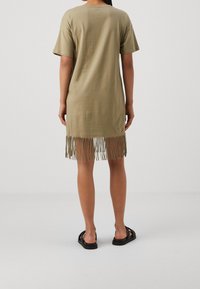 Person som står vänd bort, iklädd en kortärmad olivgrön klänning med fransad nederkant och svarta sandaler mot en enfärgad ljus bakgrund.