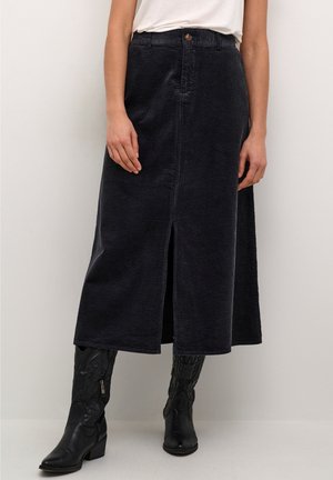 Vrouw draagt een zwarte ribfluwelen midi rok met een split aan de voorkant, een wit topje en zwarte leren mid-kuit laarzen met borduurwerk en ritsdetails.