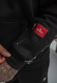 Schwarzer Kapuzenpullover mit einer Reißverschlusstasche am Ärmel, versehen mit einem roten Etikett und kontrastierenden Nähten. Weicher Stoff mit gerippten Bündchen.