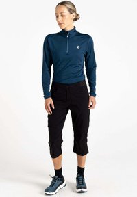 Langärmliges, dunkelblaues Oberteil mit halbem Reißverschluss und Logo, kombiniert mit schwarzen Capri-Hosen. Das Modell trägt blau-schwarze Schuhe mit schwarzen Socken.