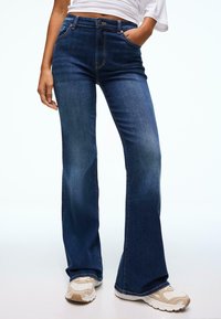 Jeans a zampa di colore blu scuro con vita alta, con tasche frontali e una leggera scoloritura. Abbinati a sneakers di colore chiaro. Tessuto in denim liscio.