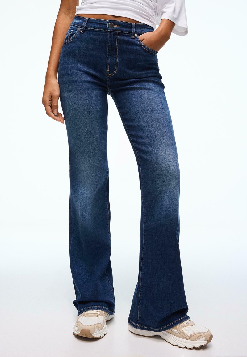 Jeans a zampa di colore blu scuro con vita alta, con tasche frontali e una leggera scoloritura. Abbinati a sneakers di colore chiaro. Tessuto in denim liscio.