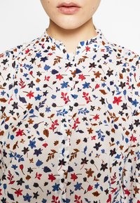 Blouse blanche légère avec un imprimé floral bleu, rouge et marron. Dotée d'un col mao et d'une fermeture boutonnée. Tissu texturé.