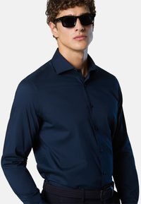 Giovane in occhiali da sole neri, camicia a bottoni blu scuro e cintura intrecciata, in piedi contro uno sfondo bianco e semplice.