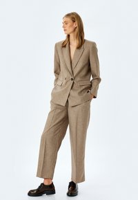 Blazer beige ajustado con solapas de pico y ribete negro, combinado con pantalones de pierna ancha a juego y zapatos negros de cordones. Textura suave.