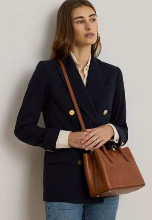 Femeie purtând un sacou negru cu două rânduri de nasturi aurii, cămașă bej, blugi albaștri, ținând o geantă maro din piele cu bretea pentru umăr.