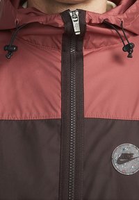 Červená a hnědá bunda na zip s kapucí, má texturovaný zip a kulatou nášivku s logem Nike se skvrnitým vzorem.