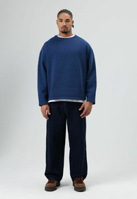Marineblauwe oversized sweatshirt met lange mouwen, gedragen over een wit overhemd, gecombineerd met donkerblauwe jeans met wijde pijpen en bruine veterlaarzen.
