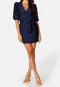 Mekko, jossa on musta ja tummansininen kietaisumalli, teksturoitu kuvio, lyhyet pömppöhihat ja solmittava vyötärö. Yhdistetty läpinäkyviin, kappalakorkoisiin sandaaleihin.