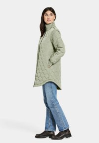 Grön quiltad kappa med hög krage, lös passform, sidfickor och rundad fåll, kombinerad med blå jeans och svarta skor.