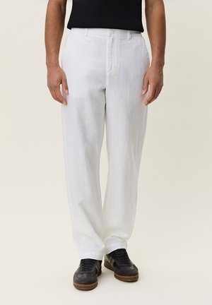 Man die losse witte broeken, een zwart overhemd en zwarte sneakers draagt, staand tegen een eenvoudige lichte achtergrond.