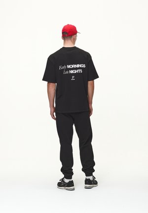 Zwart katoenen T-shirt met witte tekst "Early MORNINGS Late NIGHTS" op de achterkant, gecombineerd met zwarte sweatpants en zwarte sneakers. Rode pet achterstevoren gedragen.