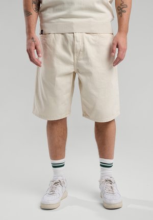 Beige katoenen short met een relaxte pasvorm, voorzien van een geribbelde tailleband, zijzakken en een gladde textuur. Gecombineerd met witte sneakers en gestreepte sokken.