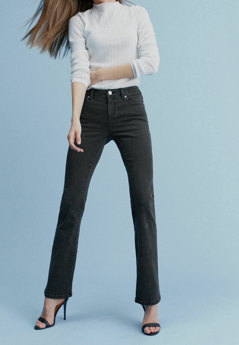 Jean en denim noir avec une coupe slim et des jambes droites, assorti à un haut blanc à manches longues côtelé. Le modèle porte des sandales noires à talons hauts.