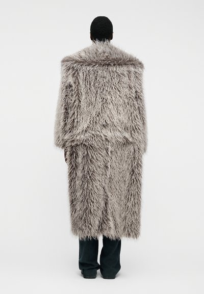 Άτομο που φοράει ένα μακρύ, χνουδωτό, ανοιχτό γκρι παλτό από γούνα, στέκεται αντικριστά με την πλάτη προς το φακό σε απλό λευκό φόντο.