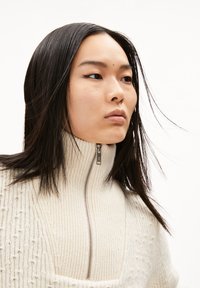 Maglione a collo alto color crema in maglia con un motivo testurizzato e una zip argentata. Finitura liscia con una vestibilità leggermente ampia.