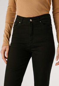 Jeans negros de talle alto con cinco bolsillos, cierre de botón metálico y detalles de costura sutiles. Combinados con una camiseta de manga larga ajustada de canalé en color beige.
