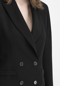 Black double-breasted blazer made of smooth fabric featuring a notched collar and six round, flat black buttons for closure.