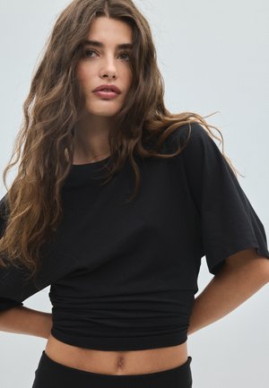 Jeune femme aux longs cheveux bruns portant un t-shirt court noir, posant avec les mains derrière le dos sur un fond clair uni.