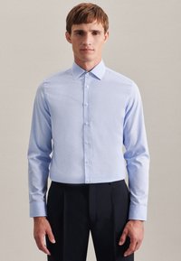 Uomo che indossa una camicia celeste a maniche lunghe con bottoni e pantaloni neri, in piedi davanti a uno sfondo beige uniforme.