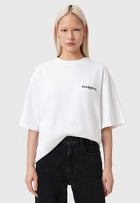 Λευκό φαρδύ t-shirt με κοντά μανίκια, διακοσμημένο με μαύρο λογότυπο στο στήθος. Συνδυάζεται με μαύρο τζιν παντελόνι ψηλόμεσο.