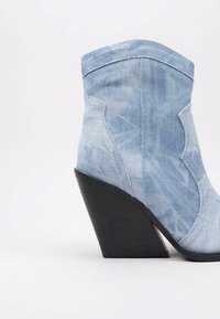 Bota de tobillo de denim con un lavado azul claro, punta afilada y un distintivo tacón de cuña. Presenta acentos cosidos y una textura suave.