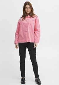 Różowa koszula dżinsowa z kołnierzykiem, zapinana na guziki, z kieszenią na piersi, w połączeniu z czarnymi dopasowanymi spodniami i błyszczącymi czarnymi butami.