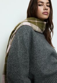 Un pull en laine grise associé à une écharpe à carreaux avec des couleurs verte, rose et beige. La texture douce et la coupe décontractée sont mises en avant.