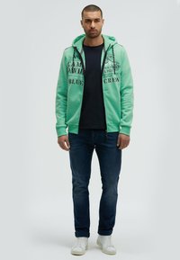 Sudadera con cremallera en verde menta con estampados gráficos, llevada sobre una camiseta azul marino, combinada con jeans azul oscuro y zapatillas blancas. Diseño sencillo, ajuste casual.