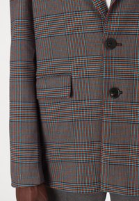 Sjekket blazer i mørkegrå med et flerfarget rutemønster med blå, oransje og svarte detaljer. Inkluderer en frontlomme og knapp.