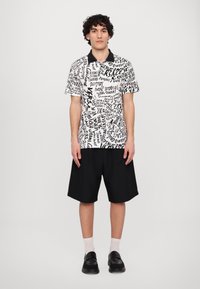 Camisa polo de manga curta em preto e branco com impressão gráfica; combinada com calções pretos. A camisa tem colarinho preto e um corte descontraído.