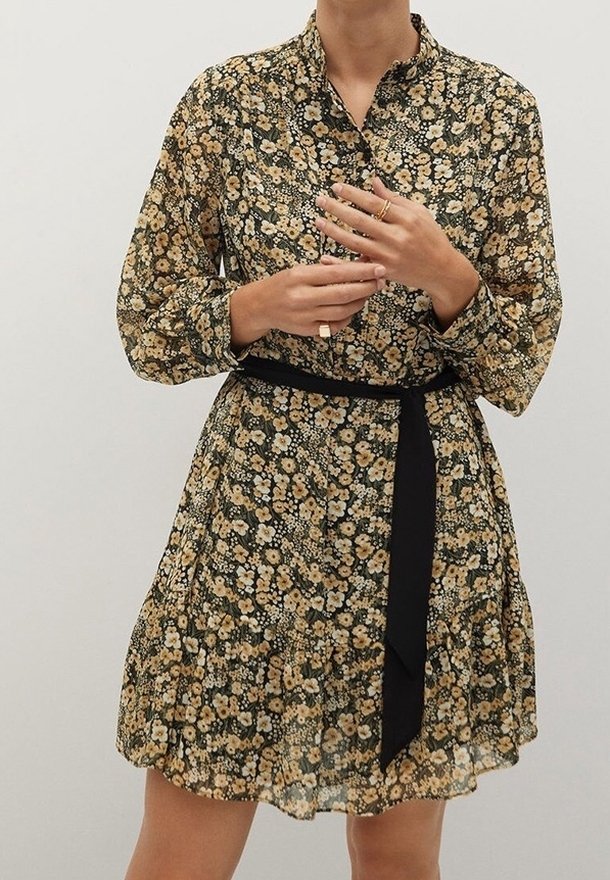 Femme portant une robe fleurie à manches longues avec une ceinture noire, ajustant des bagues à ses doigts sur un fond uni.