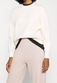 Helles beiges Sweatshirt mit einem gerippten Rundhalsausschnitt, gesticktem Logo auf der Brust und langen Ärmeln, kombiniert mit schwarzen und rosa Lagenhosen.
