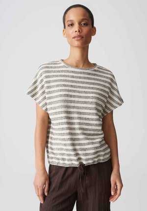 Donna dai capelli corti che indossa una maglietta a maniche corte a righe bianche e nere e pantaloni larghi marrone scuro, in piedi davanti a uno sfondo semplice.