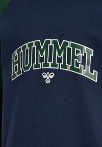 Granatowy sweter z zielonymi rękawami raglanowymi, z napisem "HUMMEL" w wyraźnych białych i zielonych literach, pod tekstem znajduje się białe logo.