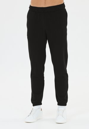 Sorte joggingbukser med elastisk talje og manchetter båret med hvide sokker og hvide sneakers på en stående person mod hvid baggrund.