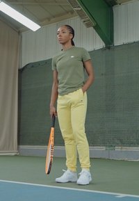 Utøver i en grønn t-skjorte og gule shorts holder en oransje tennisracket. Innendørs tennismiljø med grønt gulv og nettbakgrunn.