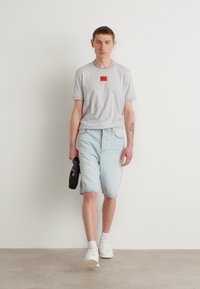 Maglietta grigia chiara a maniche corte con un logo rettangolare rosso, abbinata a pantaloni shorts in denim azzurri e sneaker bianche. Design semplice e vestibilità casual.